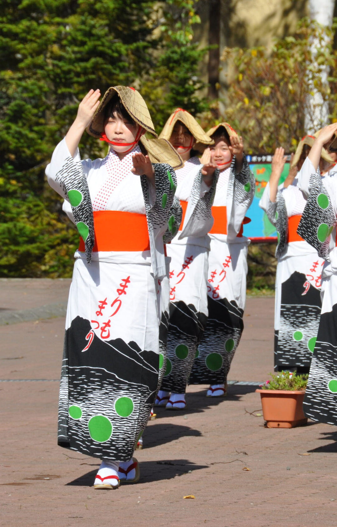 Women dressed in traditional Japanese attire performing a dance at the Marimo Matsuri festival in Lake Akan, Japan. Their kimonos feature Marimo moss ball designs with bright green circles, red sashes, and black wave patterns, and they wear woven straw hats while raising their hands in a ceremonial gesture.