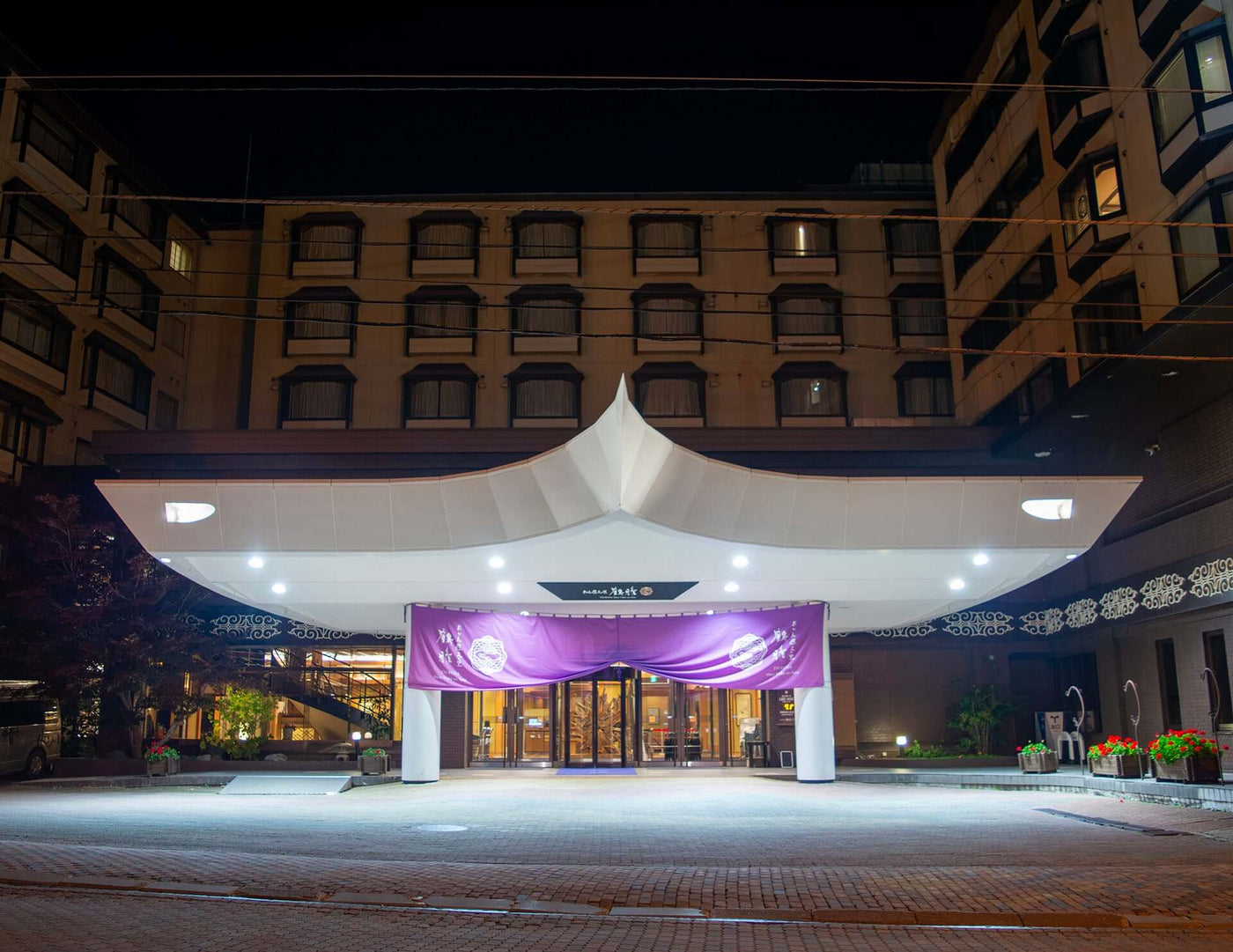 Front entrance of Akan Yuku no Sato Tsuruga, a hotel and ryokan at Lake Akan, Japan, illuminated at night with traditional purple noren curtains and modern architectural canopy.