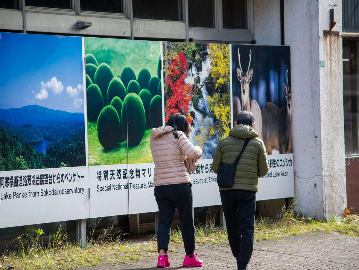 Visitors in Akanko Onsen, the lakeside town at the base of Lake Akan in Hokkaido, Japan, looking at a colorful outdoor sign featuring Marimo moss balls, a designated Special National Treasure.