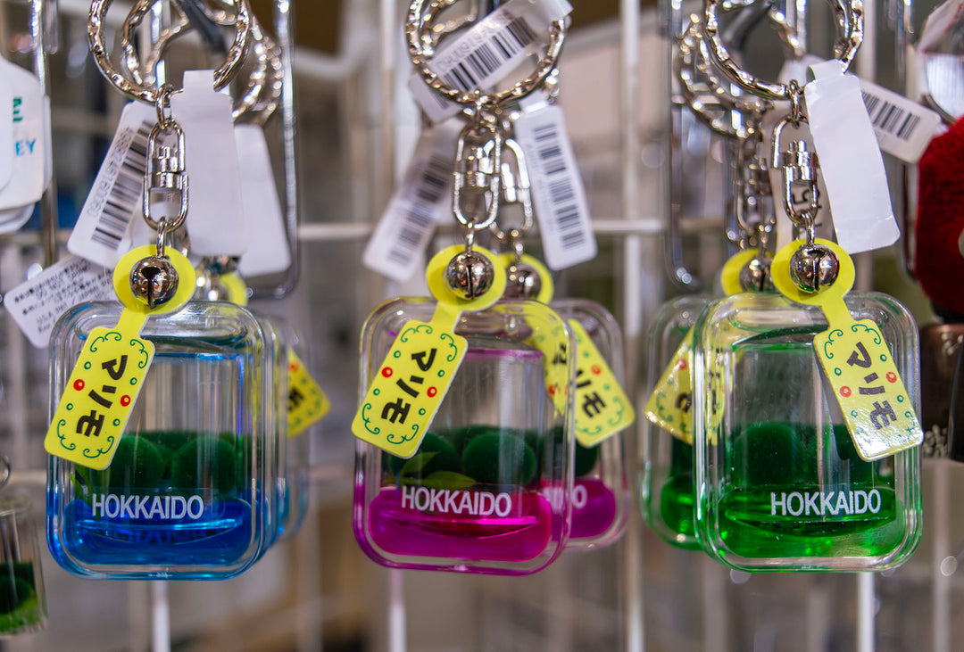 Close-up of authentic faux marimo moss ball keychains from Akanko Village in Hokkaido, Japan, featuring two green marimo floating in colored liquid inside clear acrylic cases labeled “Hokkaido,” with yellow tags and small bells attached.