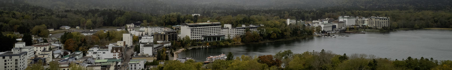 Aerial view of Lake Akan and the hot spring town of Akanko Onsen in Hokkaido, Japan, with a vibrant rainbow arching over the misty mountains — home of the famous Marimo moss balls.