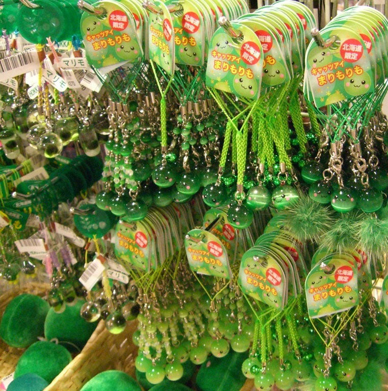 Display rack filled with colorful Marimo-themed keychains and souvenirs in various shades of green, featuring cute cartoon Marimo characters, beads, and fuzzy charms. These Hokkaido-exclusive items are popular gifts and collectibles associated with the Marimo Moss Ball and the Marimo Festival in Japan.