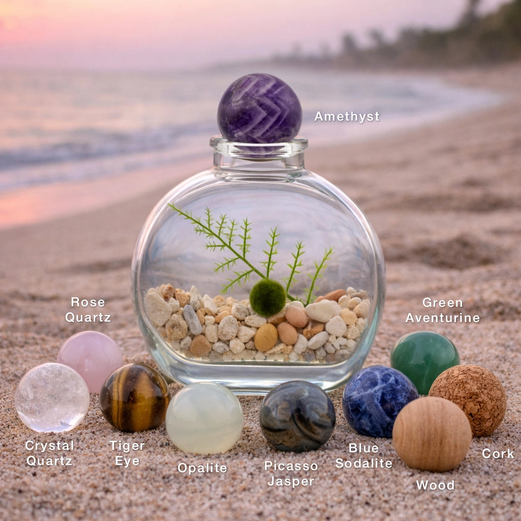 Round gemstone bottle topper spheres in the sand beside a glass marimo moss ball terrarium, including quartz, rose quartz, tiger eye, amethyst, opalite, green aventurine, sodalite, Picasso jasper, wood, and cork spheres from MossBallPets.