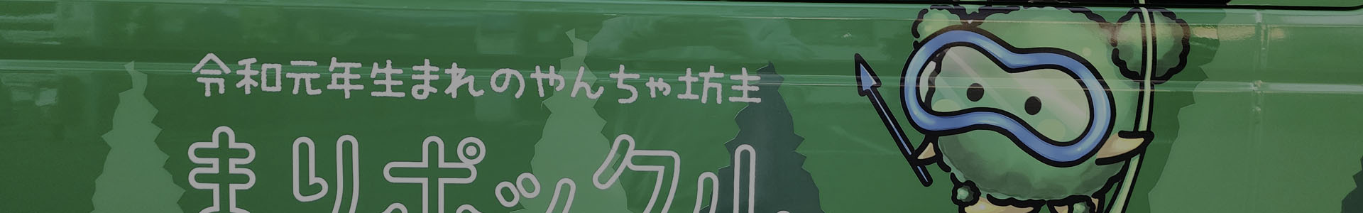 Marimo cartoon mascot character holding a lotus umbrella and snorkel gear on the side of a green bus in Kushiro, Japan — celebrating the iconic moss ball of Lake Akan.